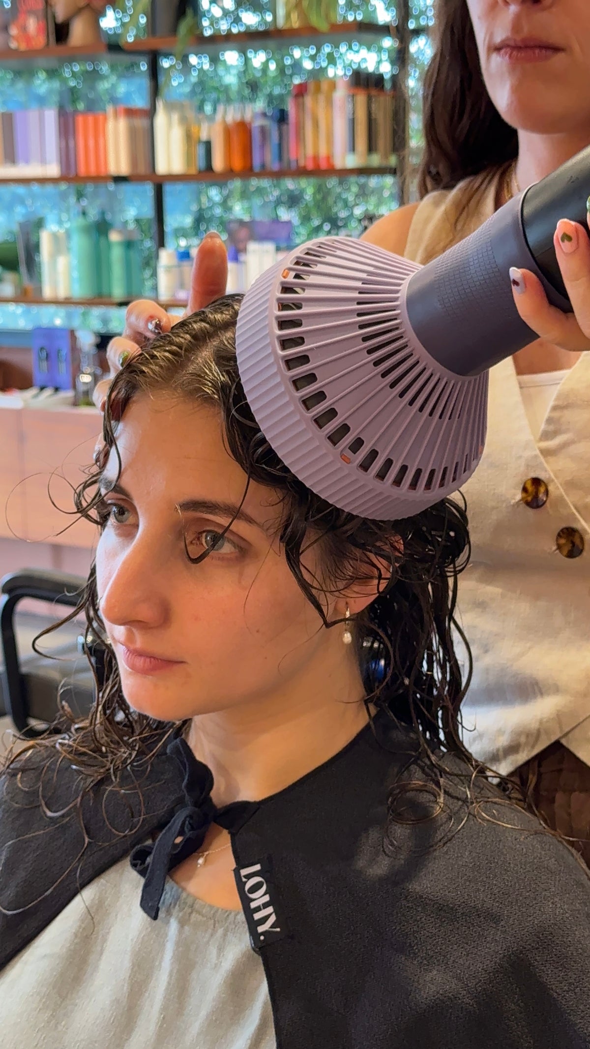 A woman in a salon setting has wet, curly dark hair while a stylist uses a handheld hair dryer with a large lavender-purple concentrator attachment. The salon interior with shelves and decor is blurred in the background.