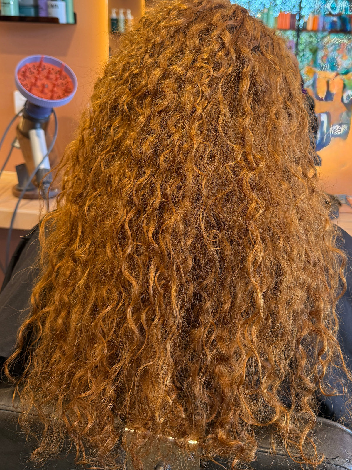 Long, voluminous curly hair in a warm coppery auburn shade, shown from the back on a person seated in a salon chair with salon equipment visible in the background. Messy, before. 