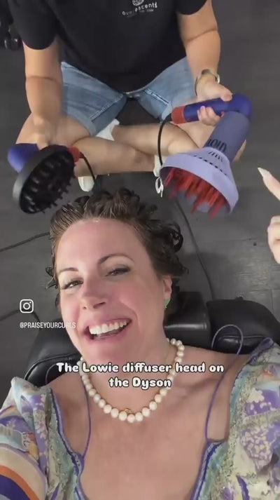 Woman laying down in a salon chair, having her hair dried by two diffusers, floral dress