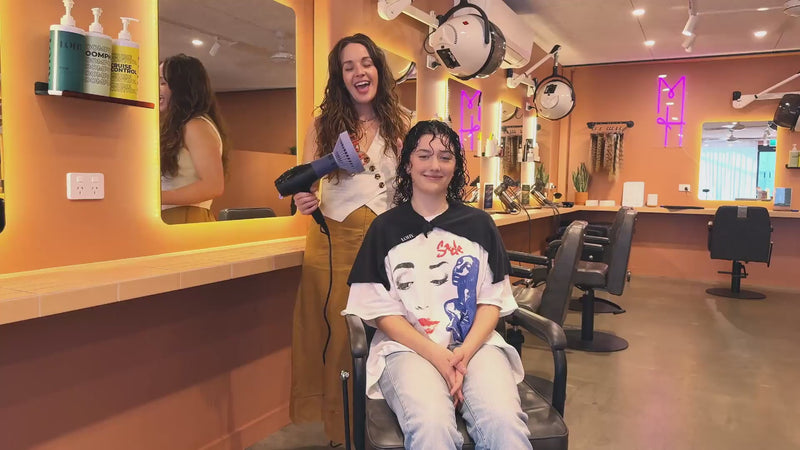 Two women in a salon, one seated and one standing, using the curl flow demonstrating how to use it, tan colours, warm tones. 